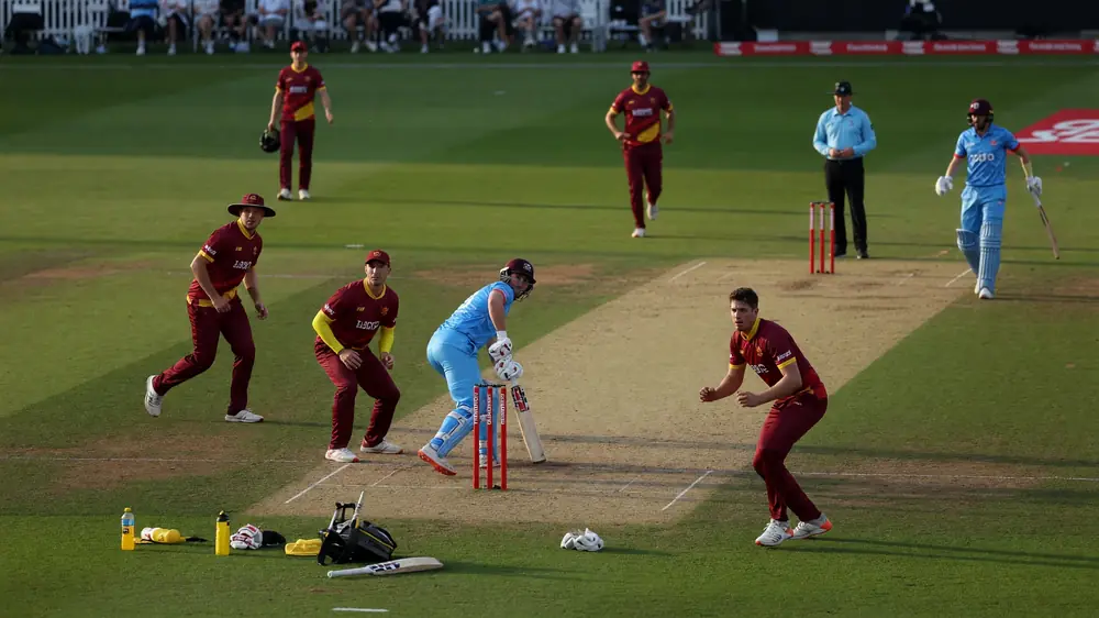 Medium shot of fielders and a bowler setting a ring on a sunlit outfield during a men’s professional cricket match, with the batter waiting at the non-striker’s end
